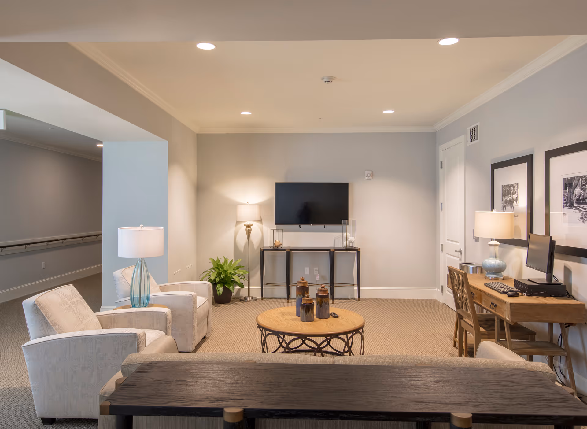 A cozy living room area in Seabrook Pointe featuring two white upholstered armchairs with a small table and lamp between them, a round wooden coffee table with decorative jars, a flat-screen TV mounted on the wall above a console table, a floor lamp, a wooden desk with a computer and chair, and two framed black and white photos on the wall.