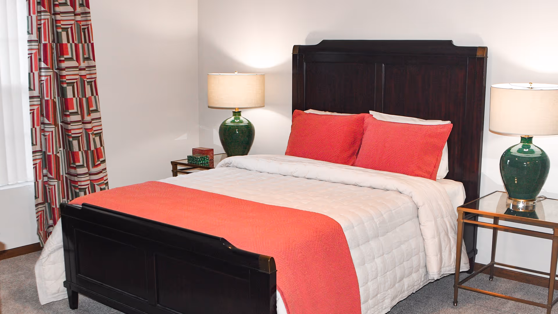 Bedroom with a dark wood bed made with a white quilt and red pillows and throw, flanked by glass-top nightstands with green lamps and patterned curtains.