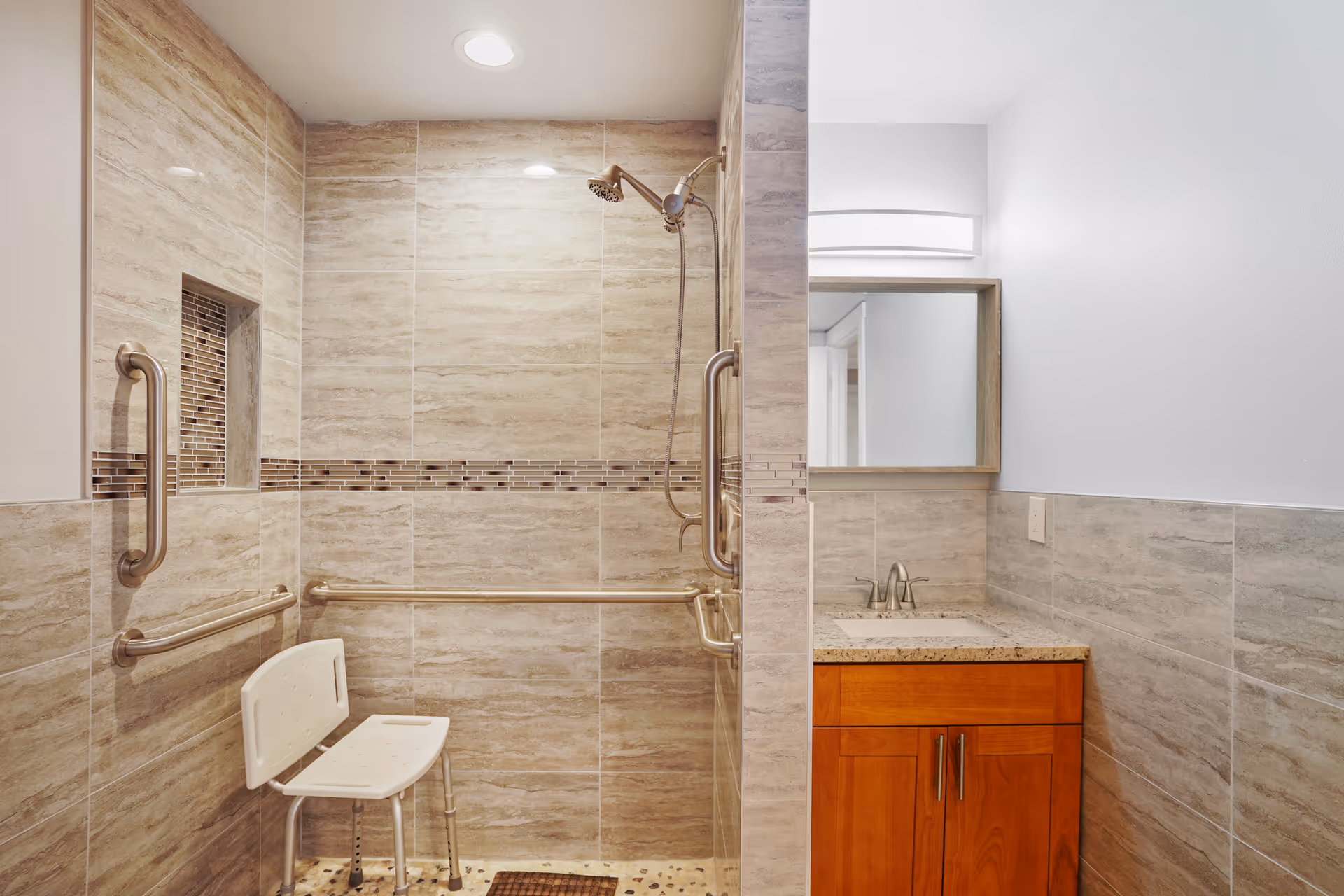A bathroom with a walk-in shower featuring beige and brown tiled walls, a white shower chair, and multiple stainless steel grab bars. To the right is a wooden vanity with a granite countertop, a sink, and a mirror above it. The lighting is bright and the space appears clean and accessible.