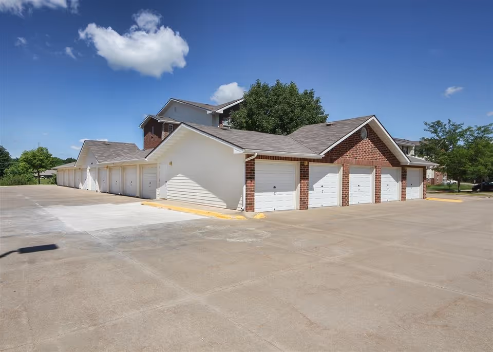A row of white garage doors attached to a brick and white siding building under a clear blue sky with a few clouds. Trees and parts of a residential building are visible in the background.