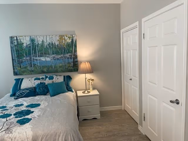 A bedroom with a bed featuring a white and teal floral bedspread and multiple pillows. Above the bed hangs a large painting of a forest scene with trees and a body of water. Next to the bed is a white nightstand with a lamp and a small decorative item. The room has light gray walls, wood flooring, and two white doors on the right side.