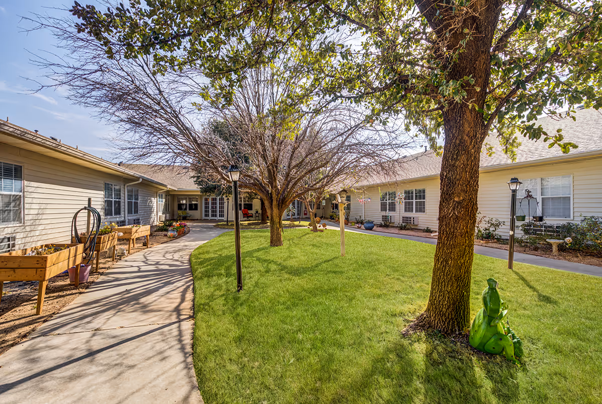 Outdoor courtyard area at Marcy Place featuring a curved concrete walkway, green grass, several trees including one with green leaves and others without leaves, garden beds with plants, decorative sculptures, and beige single-story buildings with multiple windows surrounding the courtyard.