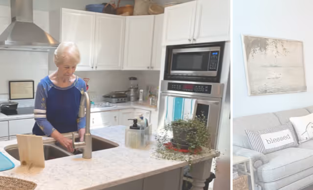 An elderly woman washing her hands at a kitchen sink in a bright, modern kitchen with white cabinets, stainless steel appliances, and a marble countertop. A small potted plant and soap dispensers are on the counter. To the right, part of a living room with a gray couch, decorative pillows, and a wall painting is visible.