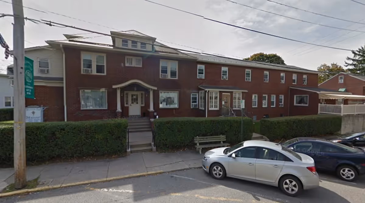 Exterior front view of a two-story brick building with multiple windows and two main entrances. There are trimmed hedges along the front and a sidewalk with parked cars in front of the building. A green banner is attached to a utility pole on the left side.