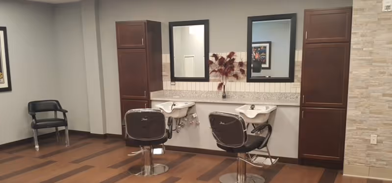 A salon corner with two styling chairs positioned at washbasins beneath mirrors, flanked by tall dark cabinets on a wood floor.