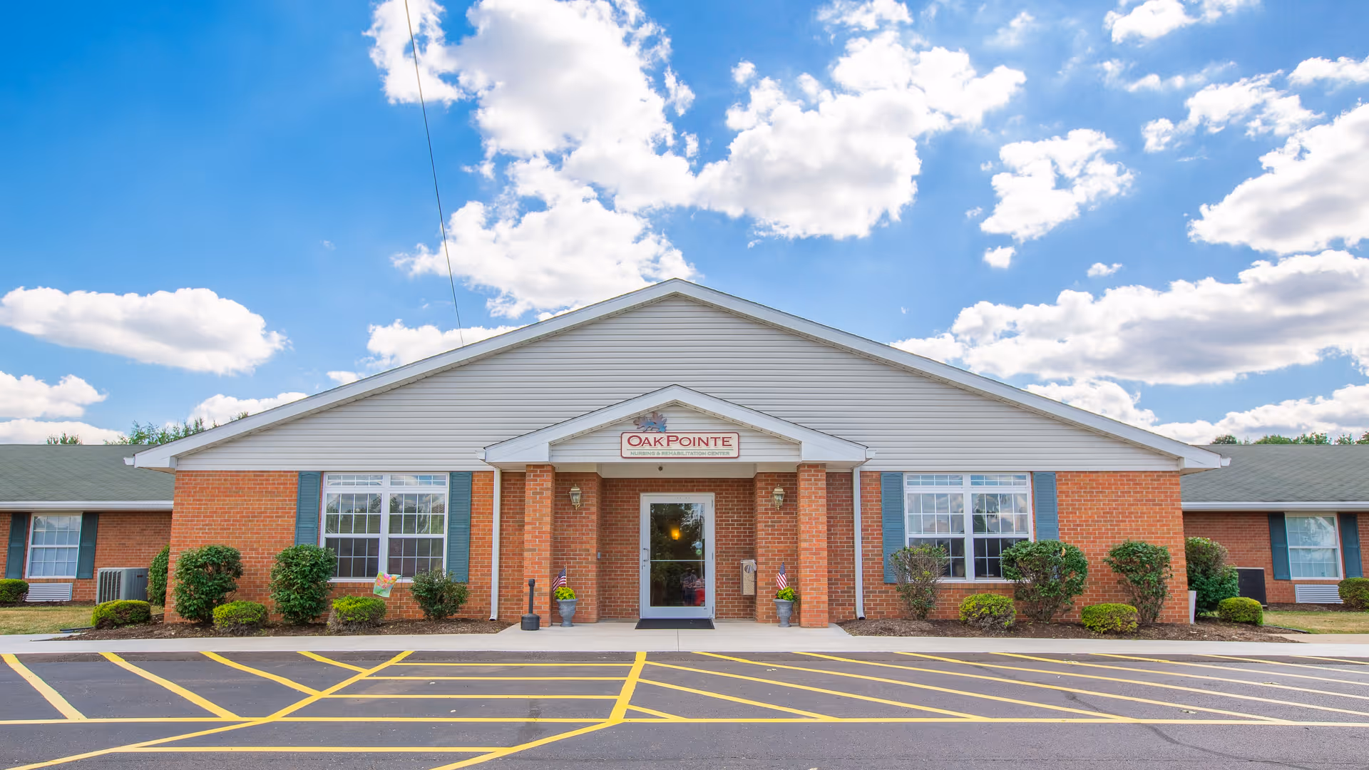 Front exterior view of Oak Pointe Nursing & Rehab Center, a single-story brick building with white siding on the upper portion, large windows with blue shutters, and a central entrance with a small covered porch. The parking lot in front has yellow painted lines and the sky is partly cloudy with blue patches.