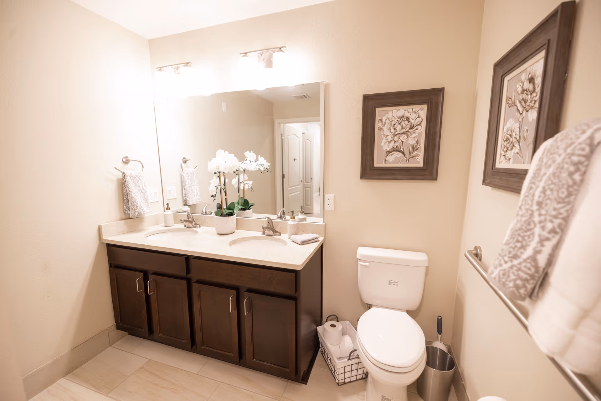 A clean and well-lit bathroom featuring a double sink vanity with dark wood cabinets, a large mirror above the sinks, and two wall-mounted light fixtures. There is a white toilet next to the vanity with a wire basket holding toilet paper rolls and a toilet brush beside it. The walls are decorated with framed floral artwork, and patterned towels hang on towel racks.