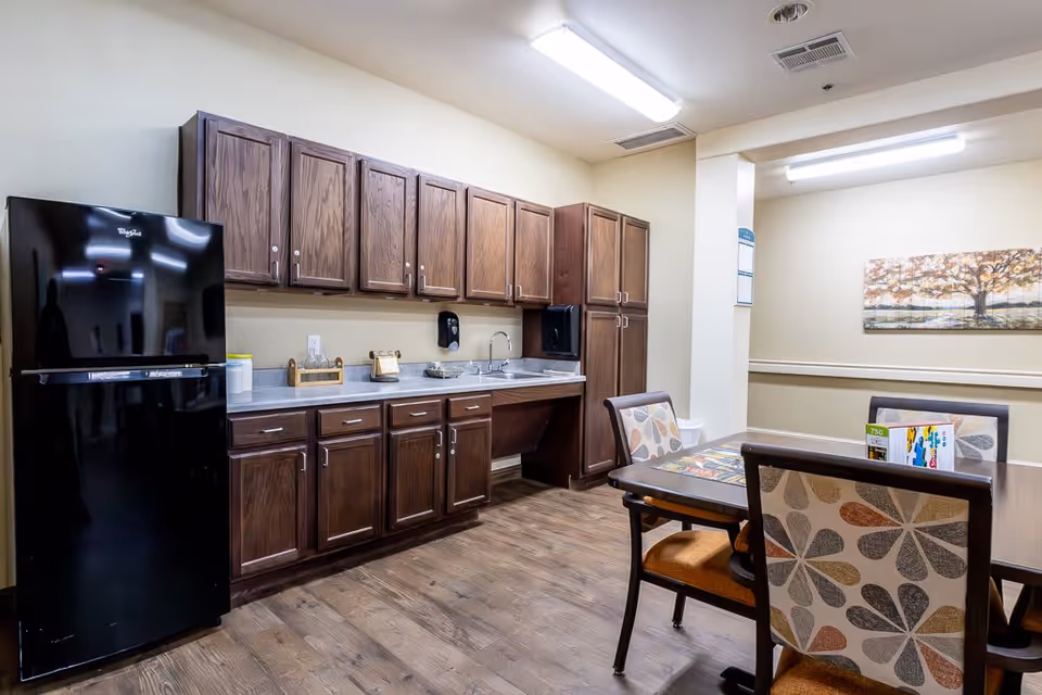 A kitchen area in an assisted living facility featuring dark wood cabinets, a black Whirlpool refrigerator, a countertop with a sink, and a dining table with four chairs that have patterned upholstery. The room has wood flooring, beige walls, fluorescent ceiling lights, and a framed picture of a tree on the wall.