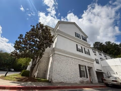White multi-story senior living building with black shutters and a parked car seen from the corner under a partly cloudy sky.