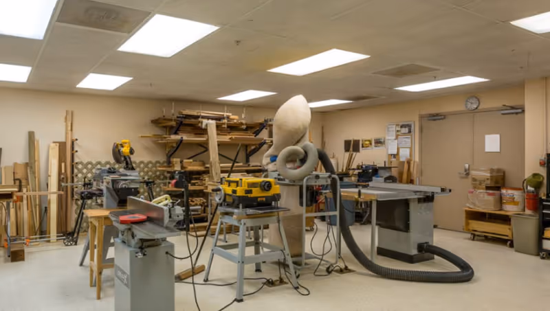 A well-lit indoor woodworking shop with saws, workbenches, stacked lumber and a dust-collection system.