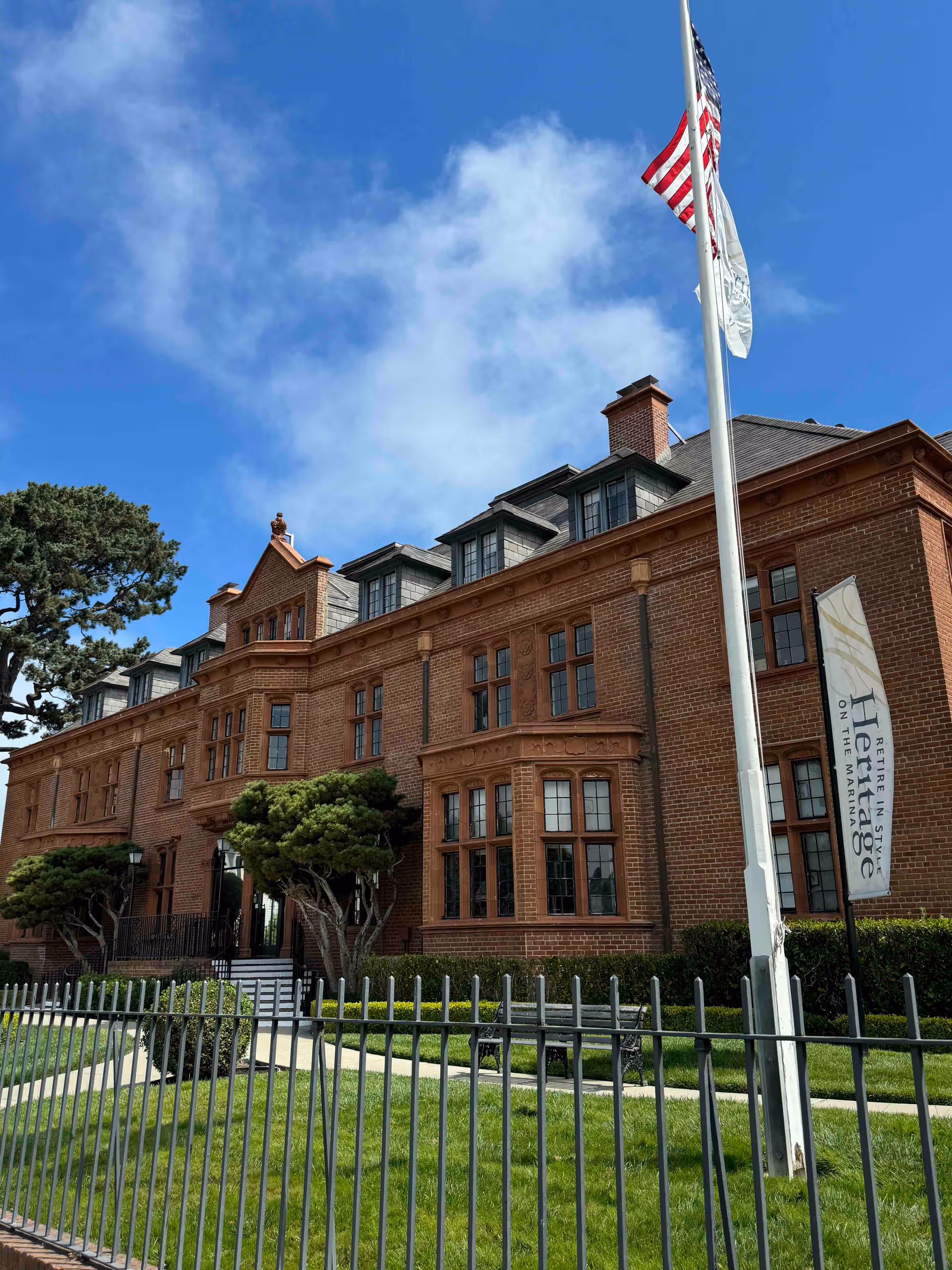 Red-brick historic-style multi-story building with dormer windows, a flagpole, and a fenced lawn in front.