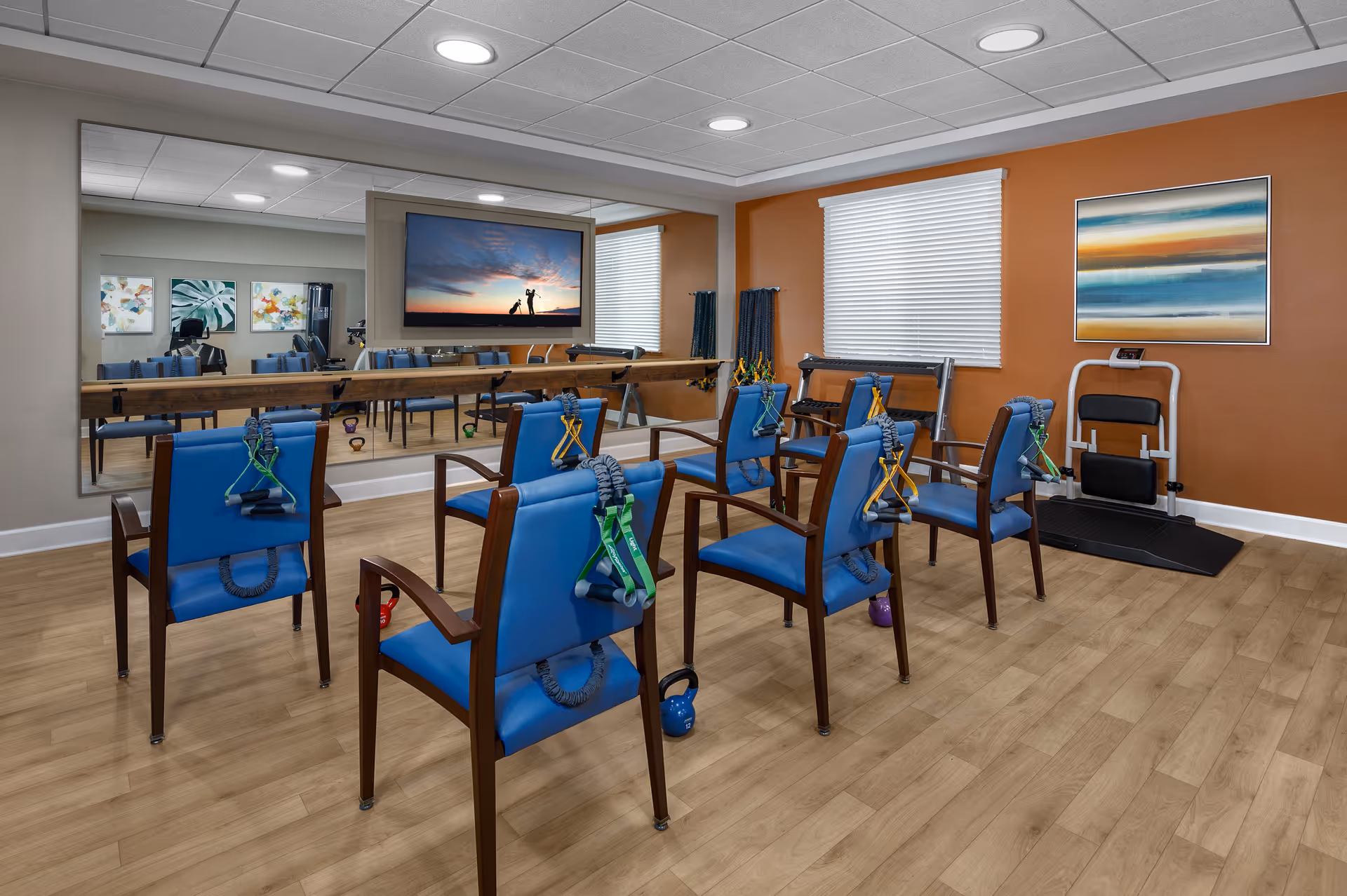 Small exercise room with blue chairs arranged in rows equipped with resistance bands and kettlebells, a wall mirror, a TV, and an exercise machine.