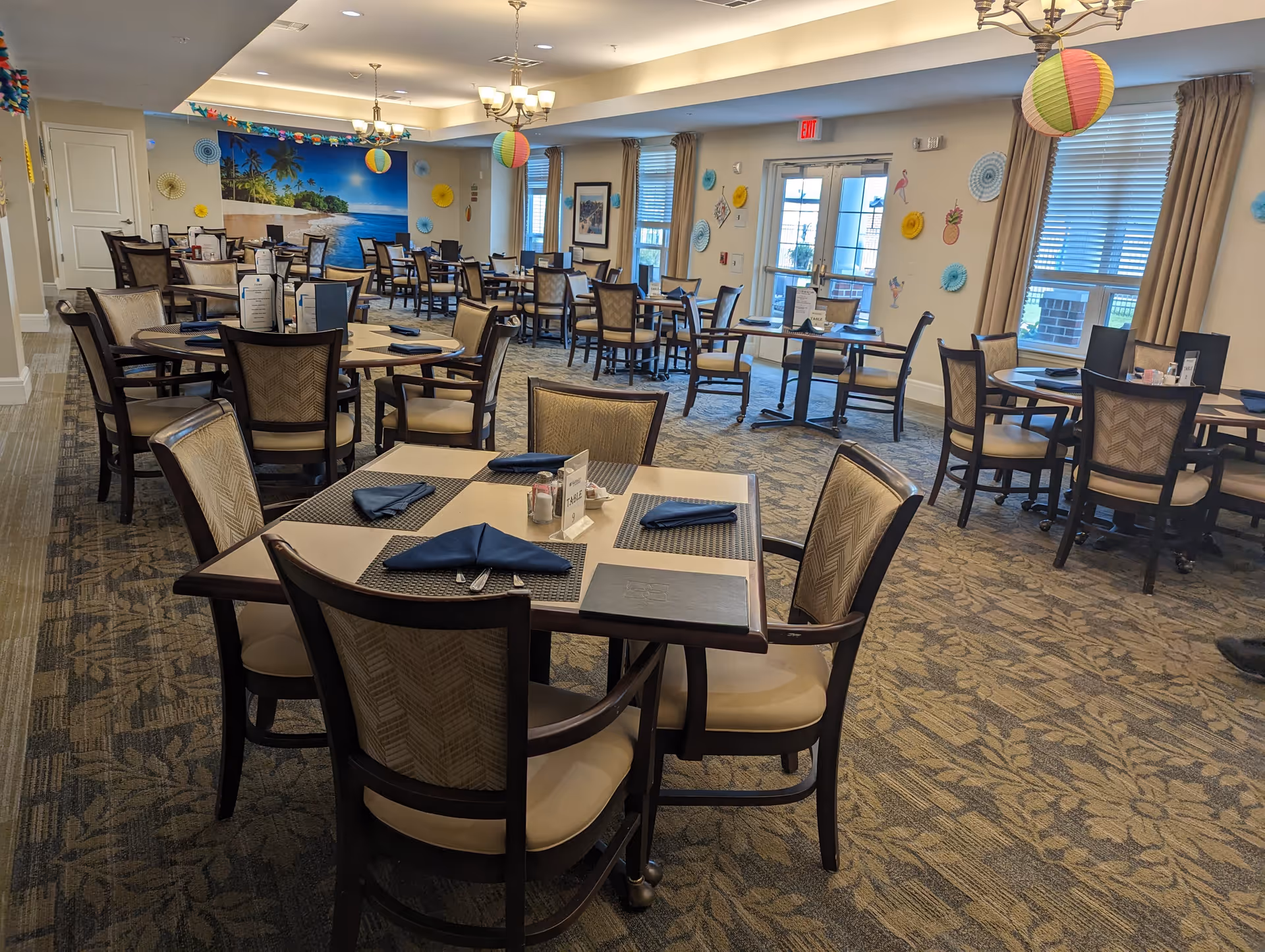 A spacious dining room with multiple tables and chairs arranged neatly. Each table is set with placemats, folded blue napkins, and menus. The room is decorated with colorful paper lanterns and wall decorations, and large windows with beige curtains allow natural light to enter. A mural of a beach scene is visible on one wall.