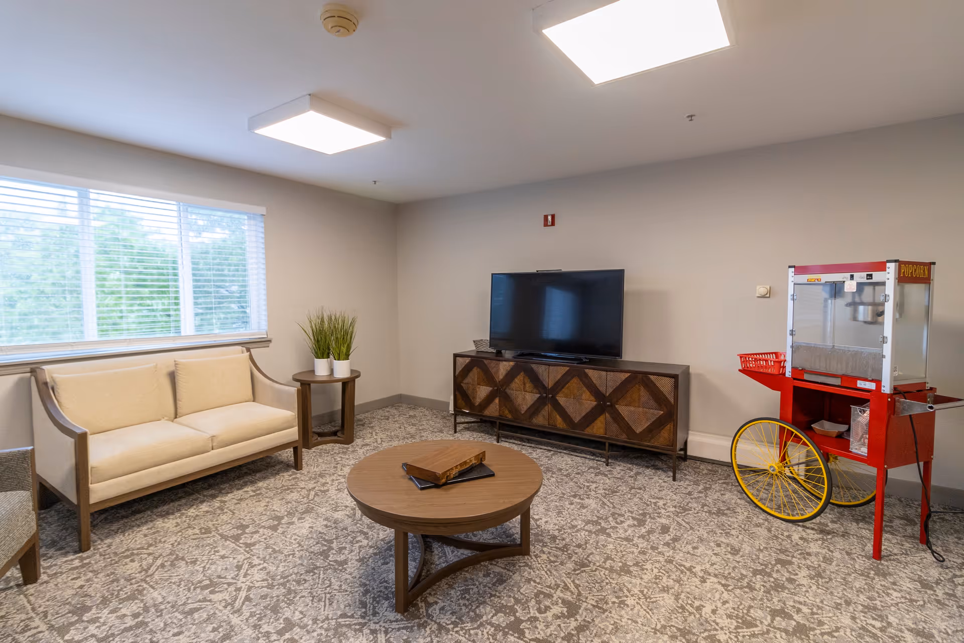 A cozy living room with a beige sofa, a round wooden coffee table with books on it, a wooden TV stand with a flat-screen television, a small side table with two potted plants, and a vintage-style red popcorn machine with yellow wheels. The room has a large window with white blinds and patterned carpet flooring.