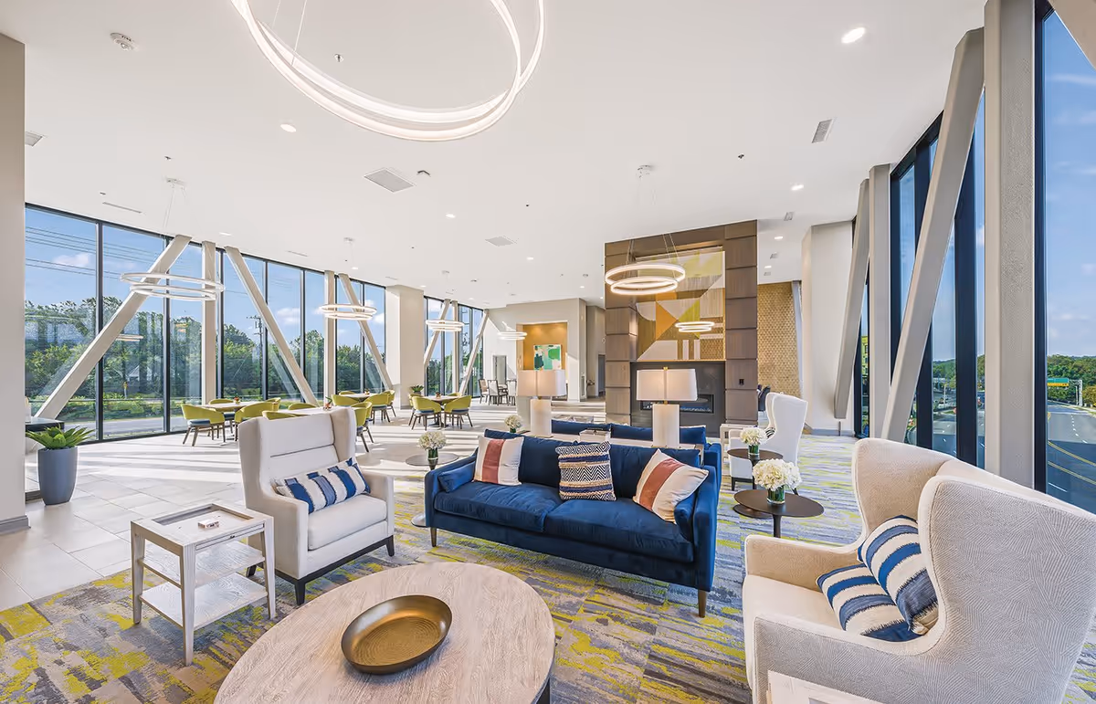 Bright modern communal lounge with sofas, armchairs, tables, and large floor-to-ceiling windows.