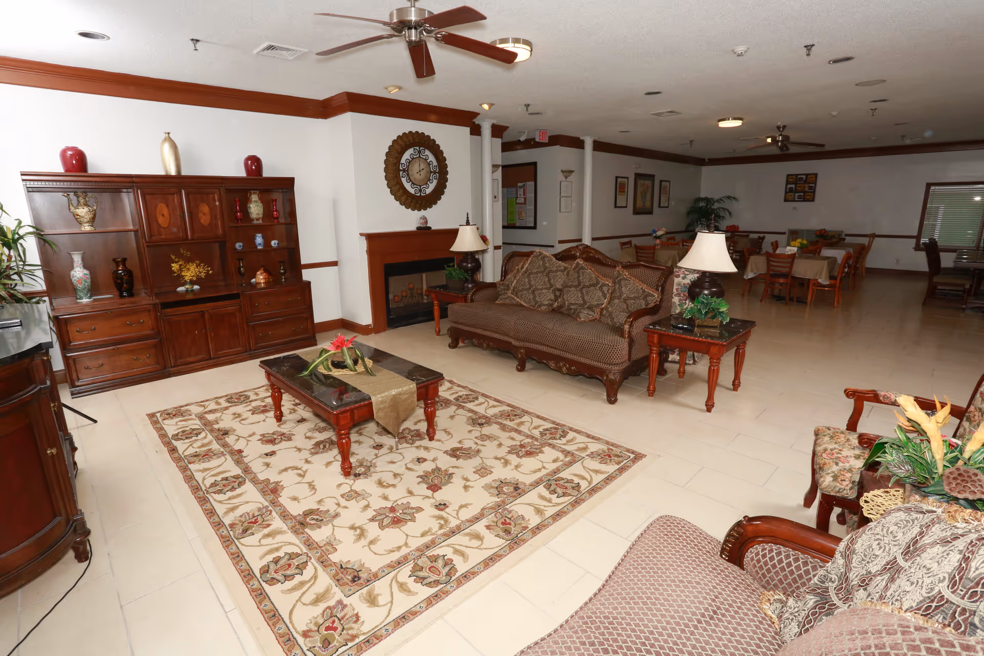 A spacious living room area in St. Francis Assisted Living Community featuring a patterned area rug, a wooden coffee table with a decorative centerpiece, a sofa with patterned cushions, two side tables with lamps, a wooden cabinet with various decorative vases, a wall clock above a fireplace, and a dining area with tables and chairs in the background.