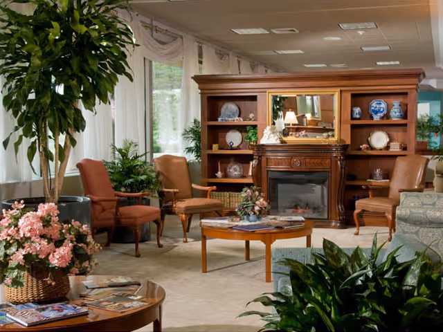 A cozy living room area in a retirement community featuring a wooden fireplace with decorative plates and vases on shelves, several upholstered chairs, a round wooden coffee table with magazines, and various green plants and flowers. Large windows with sheer curtains allow natural light to fill the space.