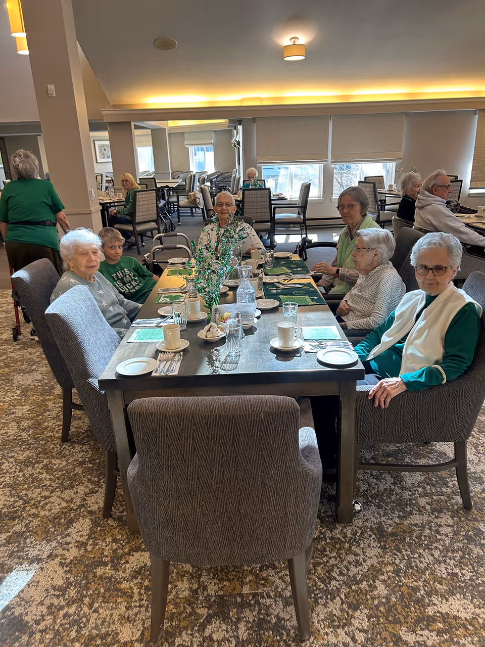 A group of elderly people and a young boy sitting around a dining table in a well-lit room with large windows. The table is set with cups, plates, utensils, and a decorative centerpiece. Other tables and chairs are visible in the background with more elderly individuals seated and engaged in conversation.