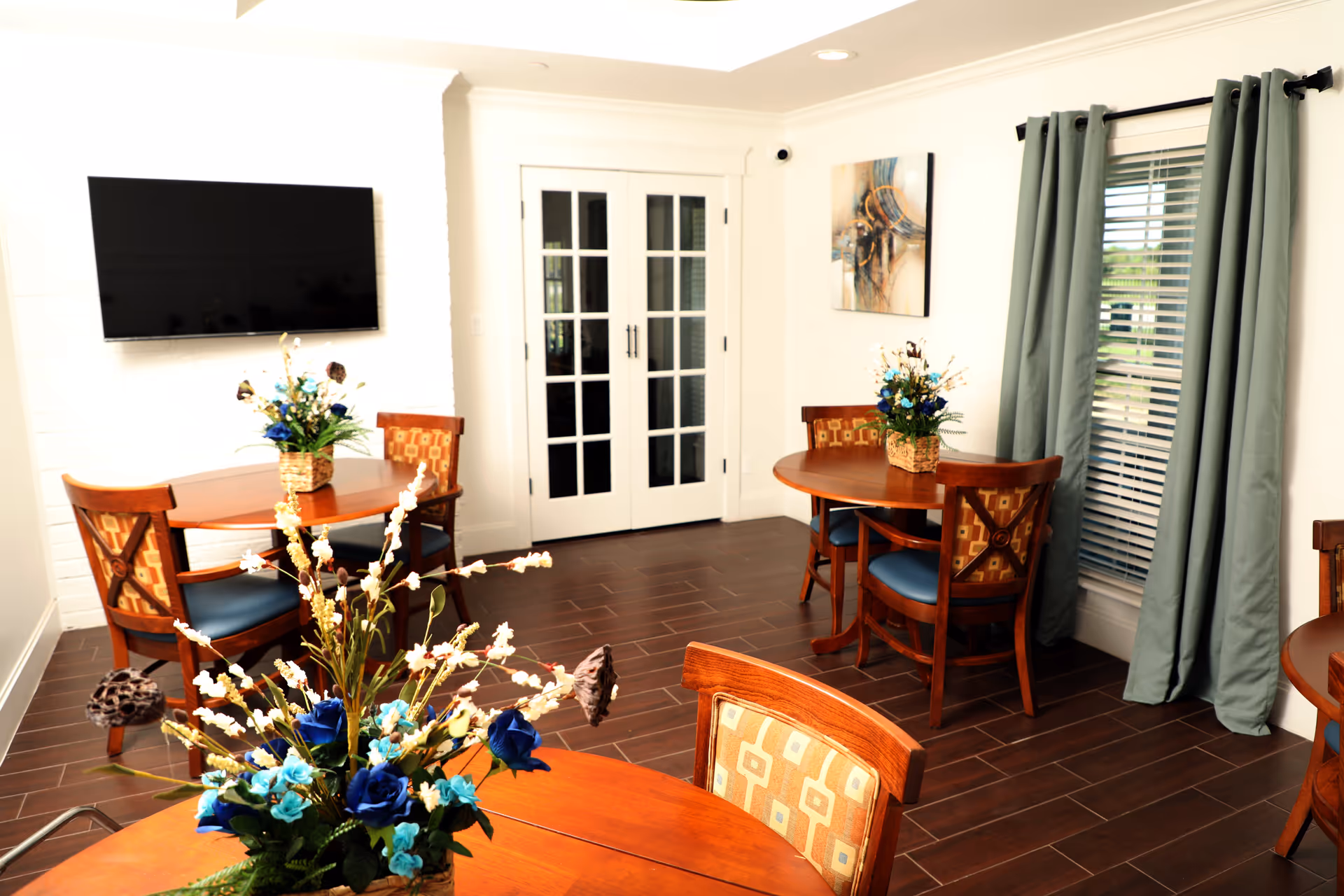 A bright and cozy dining area with wooden round tables and chairs featuring patterned cushions. Each table has a floral centerpiece with blue and white flowers. There is a wall-mounted flat-screen TV on a white brick wall, French doors with glass panes, a window with green curtains, and a piece of abstract artwork on the wall.