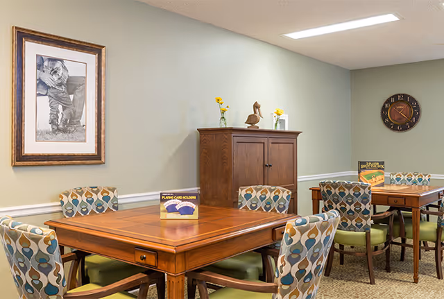 Communal dining/activity room with wooden tables, patterned chairs, a cabinet, framed wall art and a clock on a pale green wall.