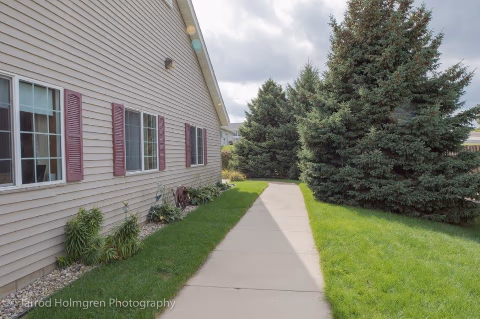 Side view of a beige building with multiple windows featuring purple shutters. A concrete walkway runs alongside the building, bordered by green grass and several tall evergreen trees. The sky is partly cloudy.