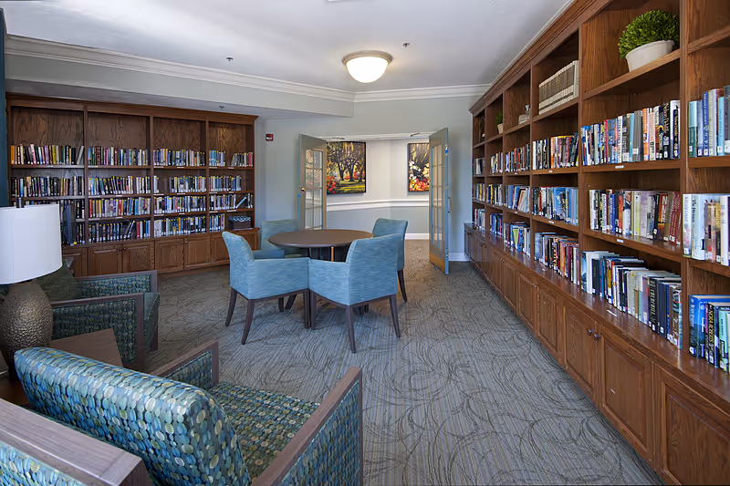 A cozy library room in a senior living facility with wooden bookshelves filled with books lining two walls. In the center, there is a round table surrounded by four blue upholstered chairs. Two patterned armchairs with a small side table and a lamp are positioned in the foreground. The room has carpeted flooring and a ceiling light fixture. Double glass doors lead to another area with framed artwork on the walls.
