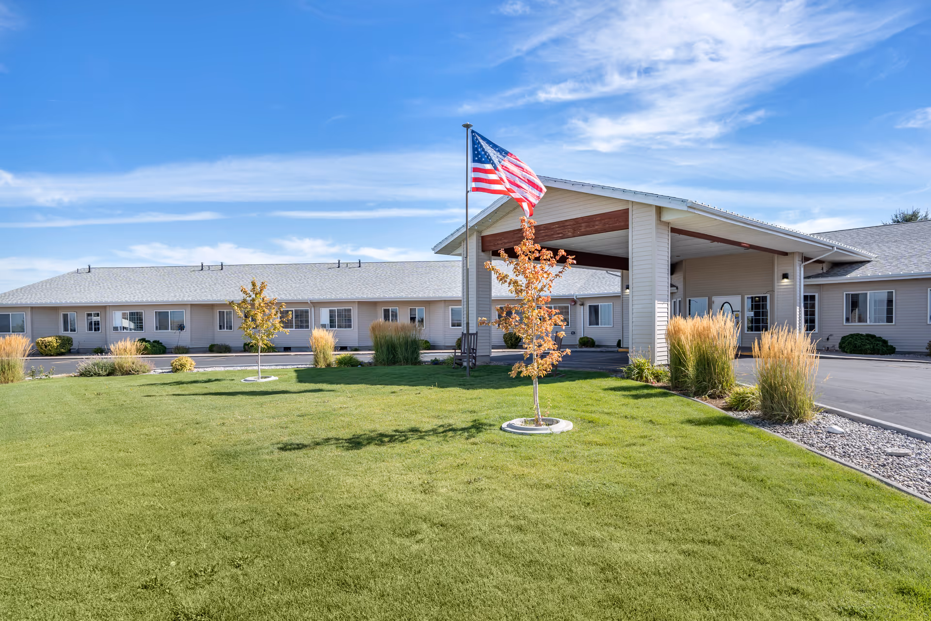 Exterior view of Brookdale Pocatello senior living facility with a large green lawn, small trees, ornamental grasses, and an American flag on a flagpole in front of the building under a blue sky with some clouds.