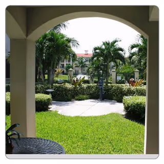 View through an arched patio opening showing a well-maintained green lawn, palm trees, shrubs, and a paved walkway with a small statue in the center. A round metal table is partially visible in the foreground.