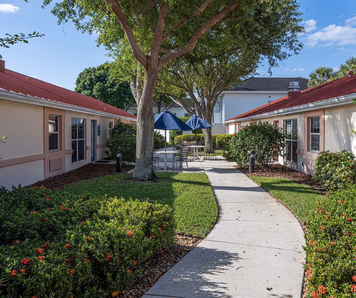 Outdoor courtyard area at Arden Courts - ProMedica Memory Care Community in West Palm Beach, featuring a paved walkway flanked by green bushes with red flowers, trees providing shade, and tables with blue umbrellas and chairs for seating.