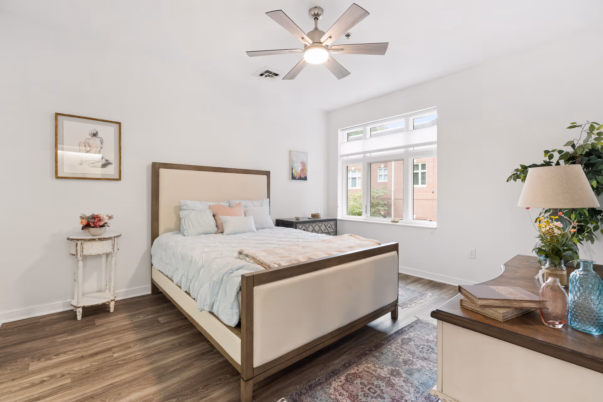 A bright bedroom with a large bed featuring a beige upholstered headboard and footboard, light blue bedding, and several pillows. The room has wooden flooring, a ceiling fan with light, a window with a view of a brick building outside, a small round side table with a flower arrangement, a dresser with decorative items including a lamp and vases, and framed artwork on the walls.