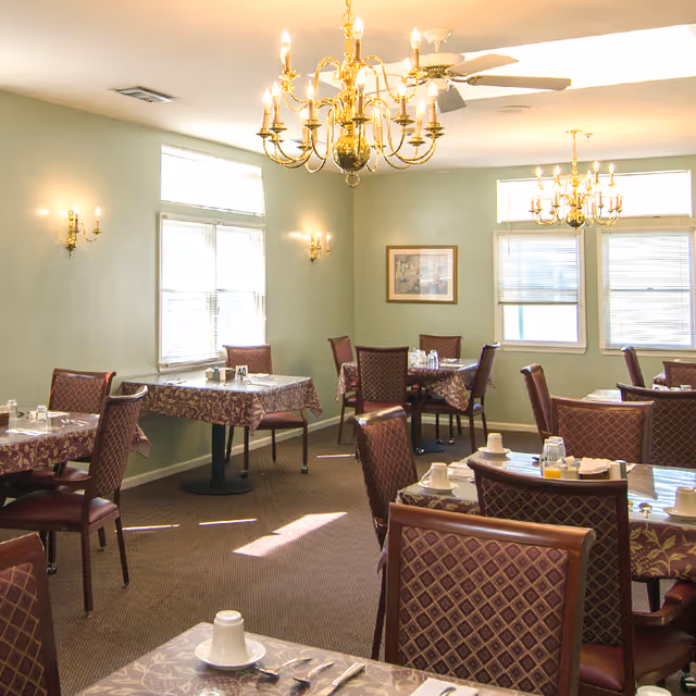 Dining room with multiple tables and chairs, chandeliers, and place settings in a senior living facility.