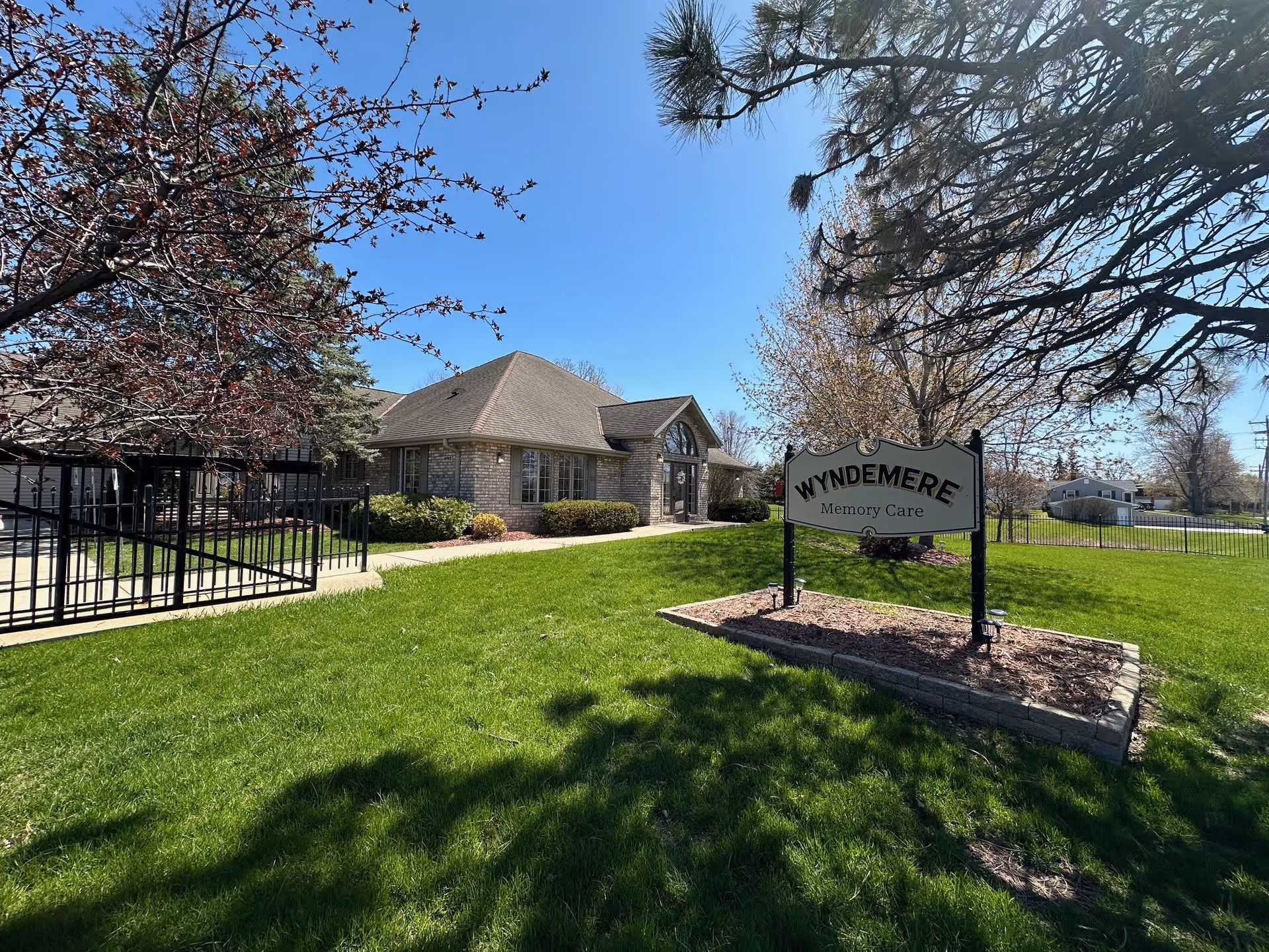 Exterior view of Wyndemere Memory Care facility on a sunny day, showing a single-story brick building with a pitched roof, surrounded by green grass, trees, and a black metal fence. A sign in the foreground reads 'Wyndemere Memory Care'.