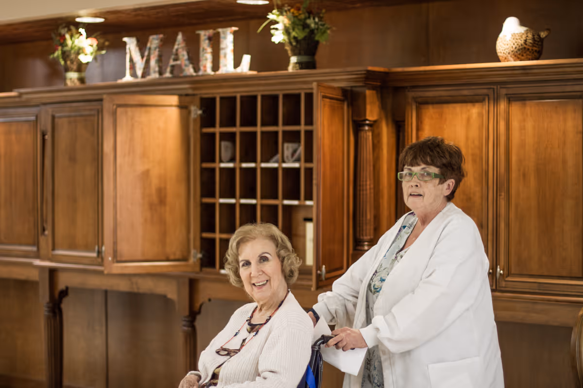 An elderly woman in a wheelchair smiling while being pushed by a caregiver in a white coat in front of a wooden mail cubby area with decorative plants and the word 'MAIL' displayed on top.