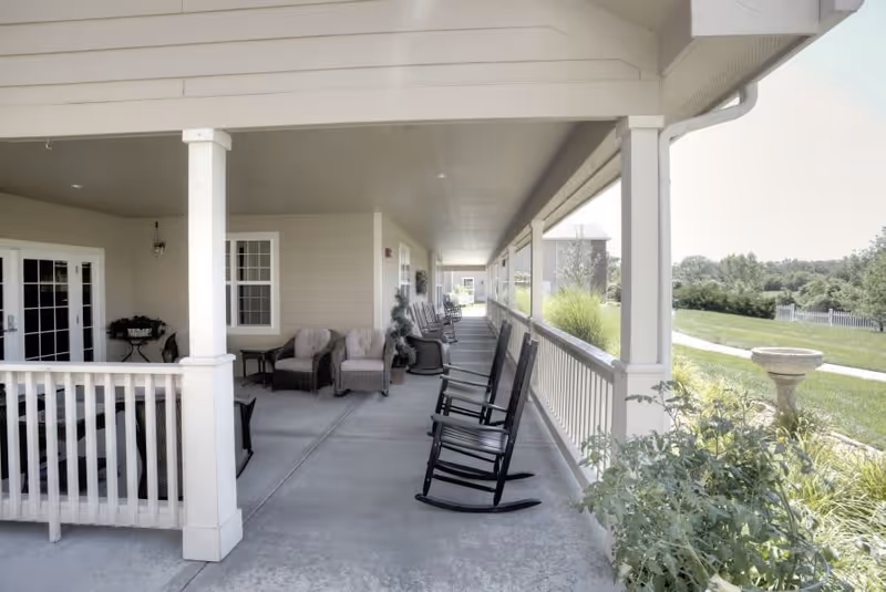 Covered outdoor porch area with several black rocking chairs lined up along the railing, cushioned armchairs and small tables near the building wall, overlooking a green lawn with trees and a birdbath.