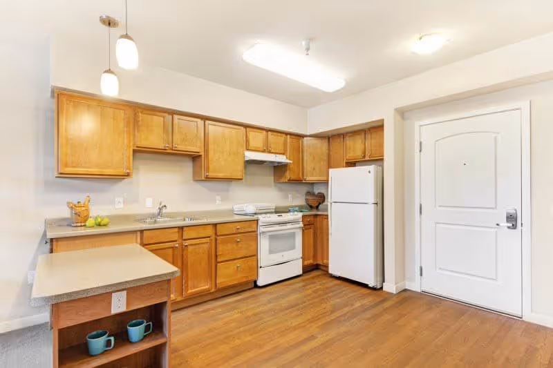 A clean kitchen with wooden cabinets, a white refrigerator, a white stove with an overhead vent, a countertop with a sink, and a small island with two blue mugs on the lower shelf. The floor is wooden, and there is a white door to the right side of the kitchen.