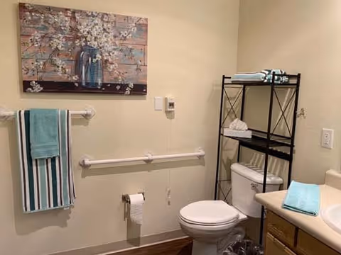 A bathroom with a toilet, a towel rack holding two towels, a black metal shelving unit with folded towels and tissue box, a sink with a blue towel on the counter, and a floral painting on the wall.