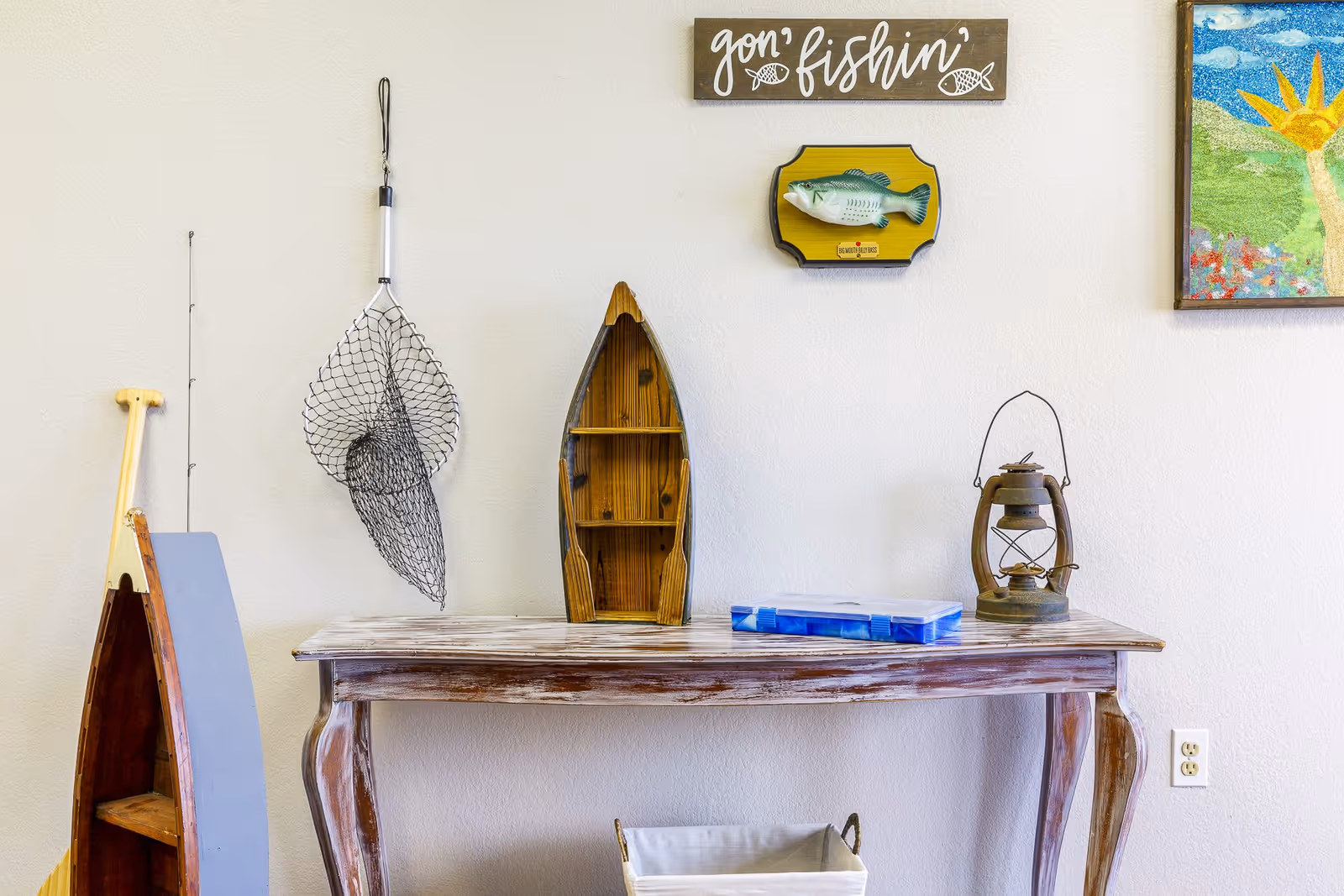 Wooden console table with nautical decor — a fishing net, small boat-shaped shelf, lantern, tackle box, and a "gon' fishin'" sign on a pale wall.