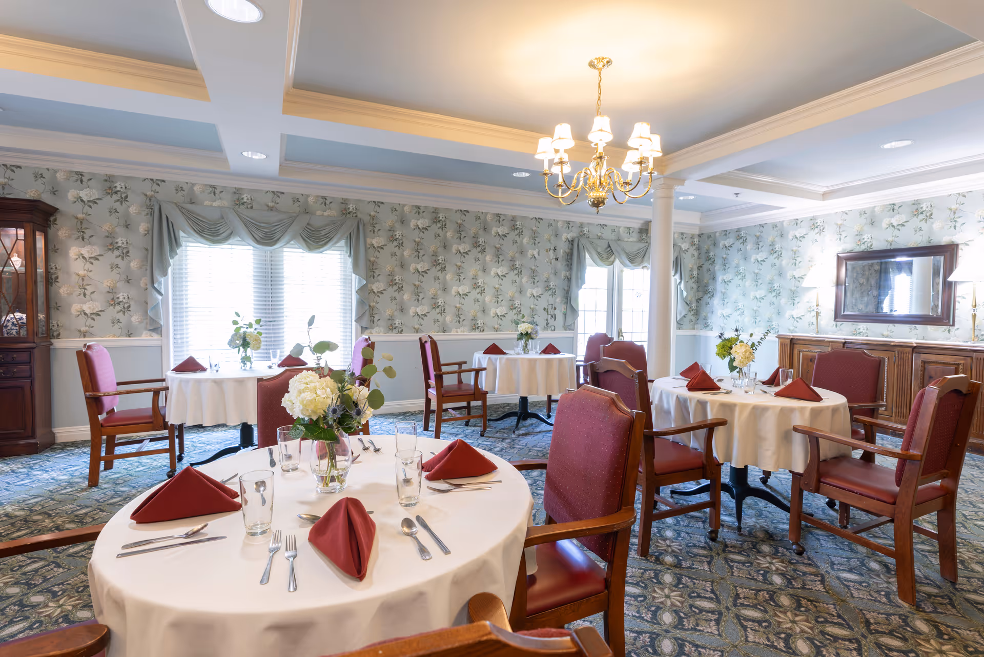 A well-lit dining room with round tables covered in white tablecloths, each set with red folded napkins, glasses, and silverware. The room features floral wallpaper, a chandelier, wooden chairs with red cushions, and a cabinet with a mirror on the wall.