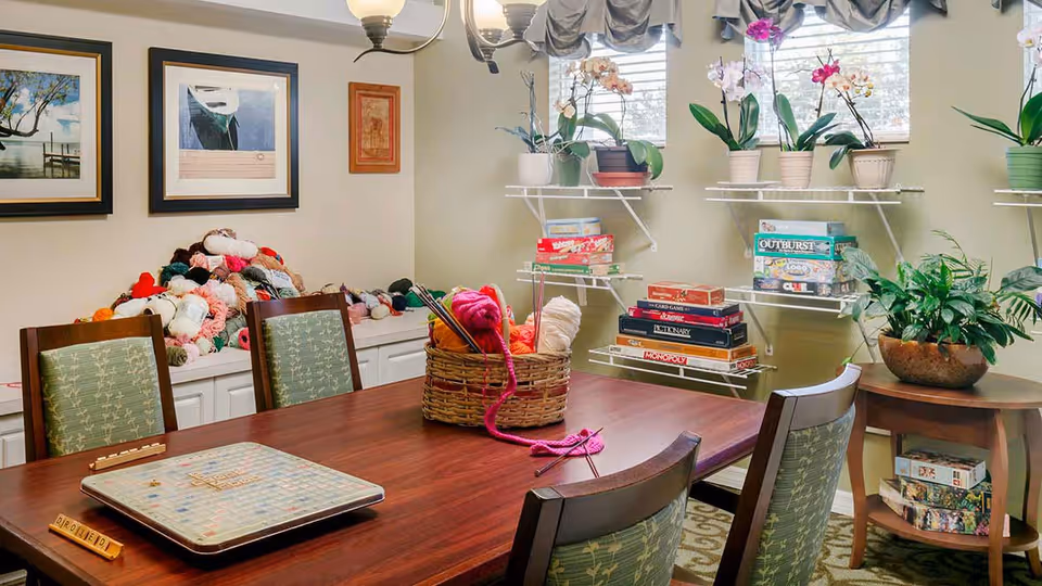 A cozy activity room with a wooden table and four chairs featuring green patterned cushions. On the table, there is a Scrabble game and a basket filled with yarn and knitting needles. Behind the table, shelves hold various board games and potted plants, while a countertop in the corner is piled with colorful yarn. The room is softly lit by a ceiling light fixture and natural light coming through three windows with decorative valances.