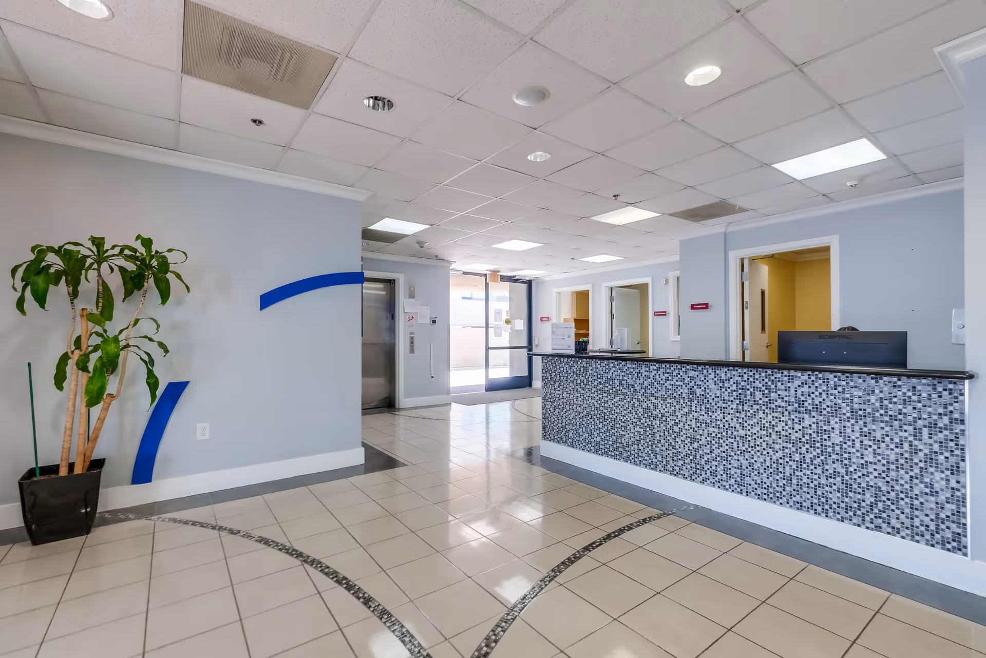 Reception area of Santa Monica Rehabilitation Center with a tiled floor, a mosaic tiled front desk, a potted plant on the left, an elevator door, and an entrance door with glass panels in the background.