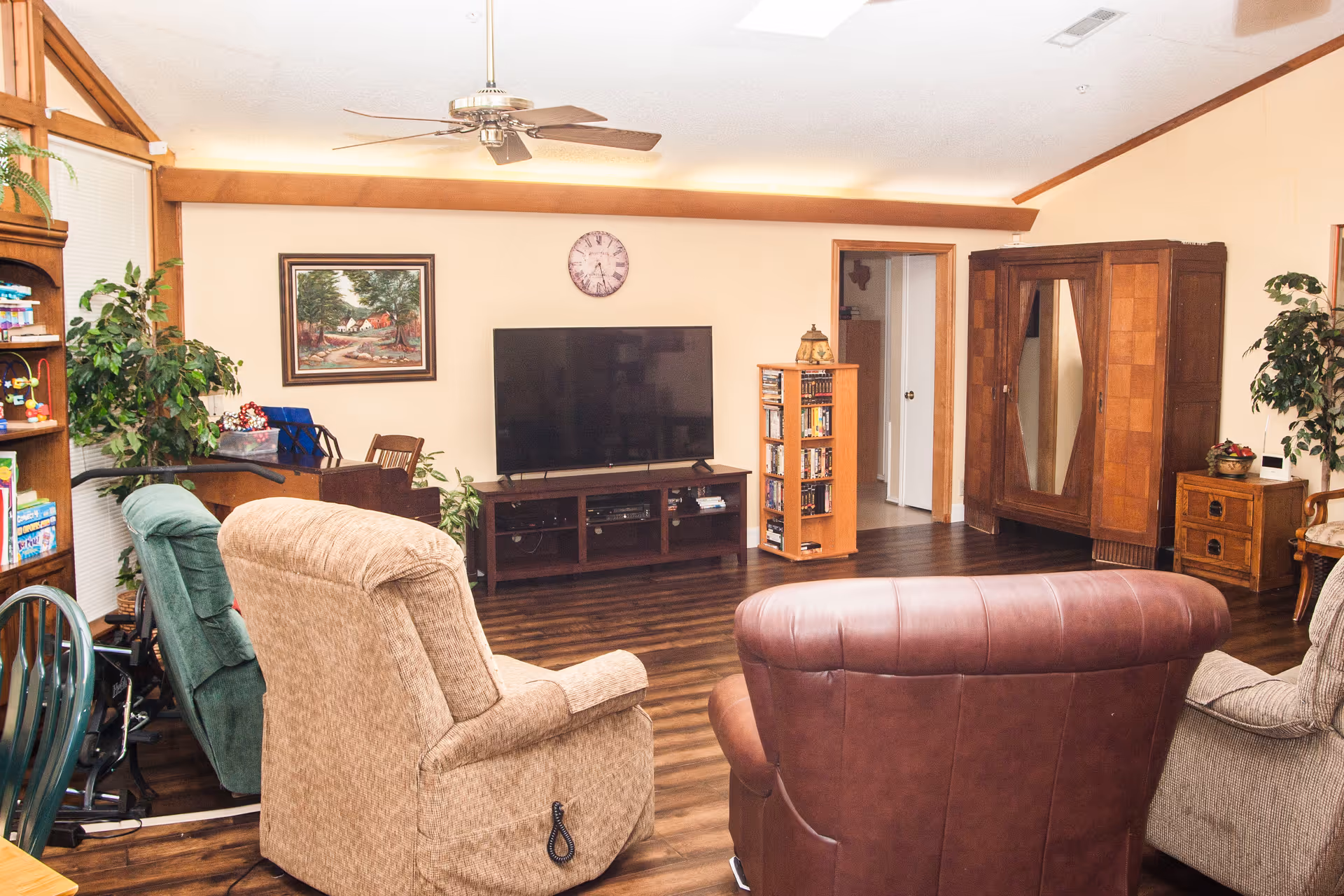 A cozy living room with a large flat-screen TV on a wooden stand, surrounded by various chairs including a beige recliner, a green chair, and a brown leather chair. The room features wooden flooring, a ceiling fan, a framed painting on the wall, a clock, and several plants. There is also a wooden cabinet and a small bookshelf with DVDs.