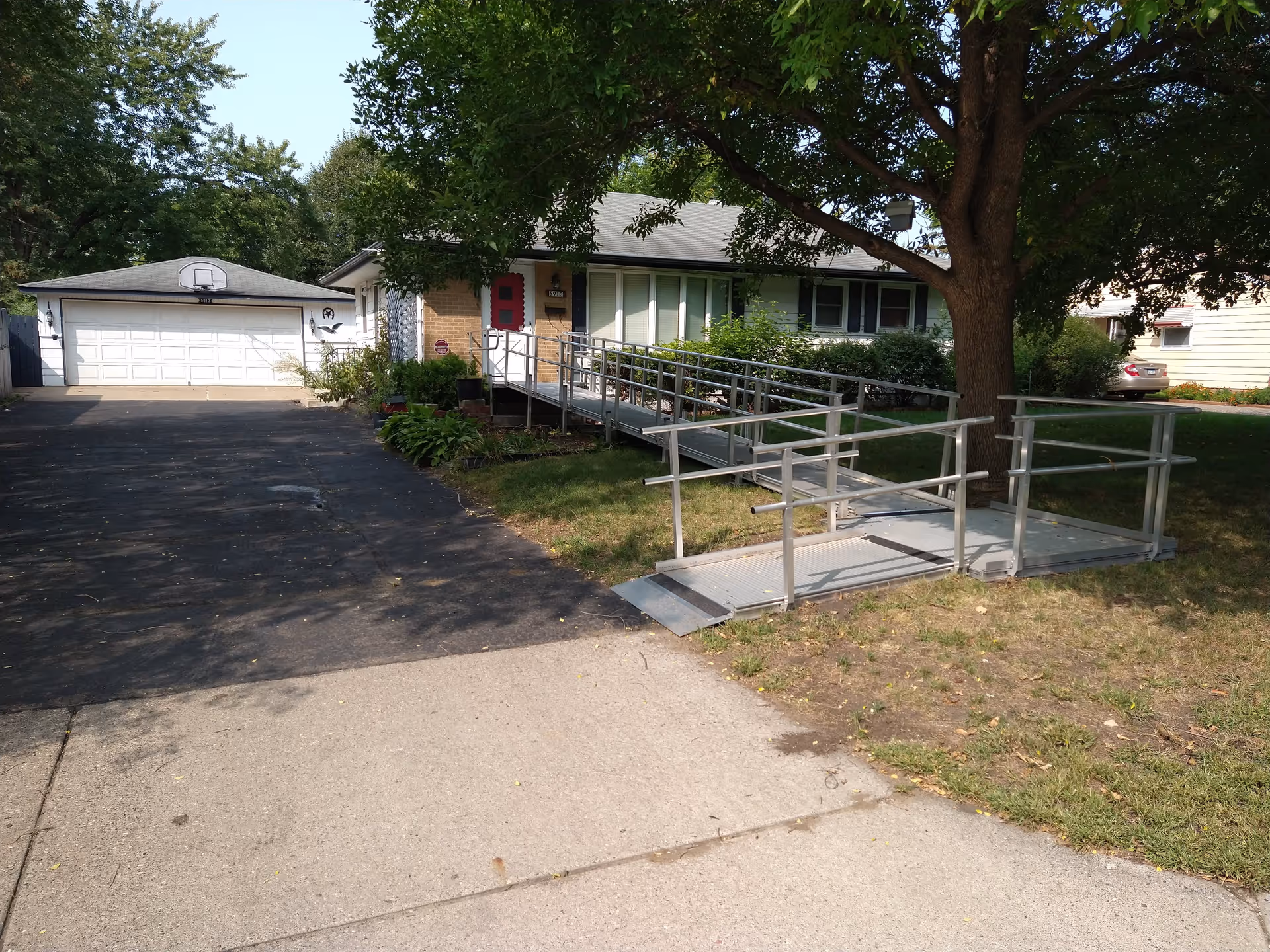 Single-story house with a wheelchair accessible metal ramp leading to the front door, a large tree providing shade, a driveway leading to a two-car garage with a basketball hoop, and surrounding greenery.