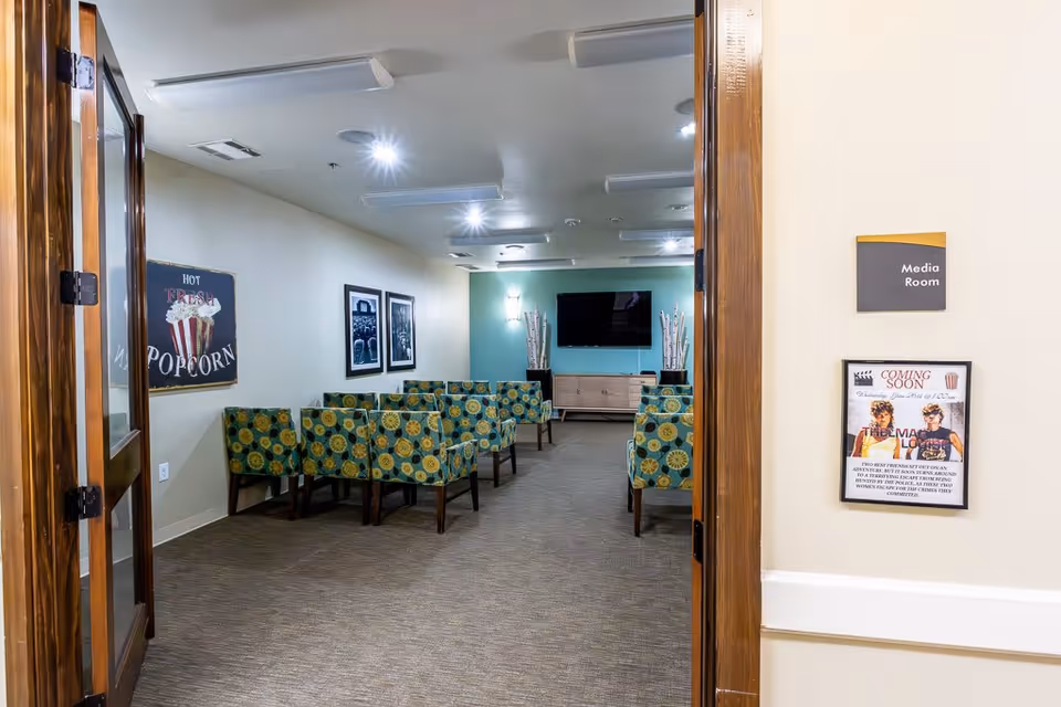 View through open double wooden doors into a media room with rows of green patterned chairs facing a wall-mounted flat screen TV. The room has beige walls with framed pictures and a sign that reads 'Hot Fresh Popcorn'. A sign outside the room says 'Media Room' and a poster below it announces a 'Coming Soon' movie event.