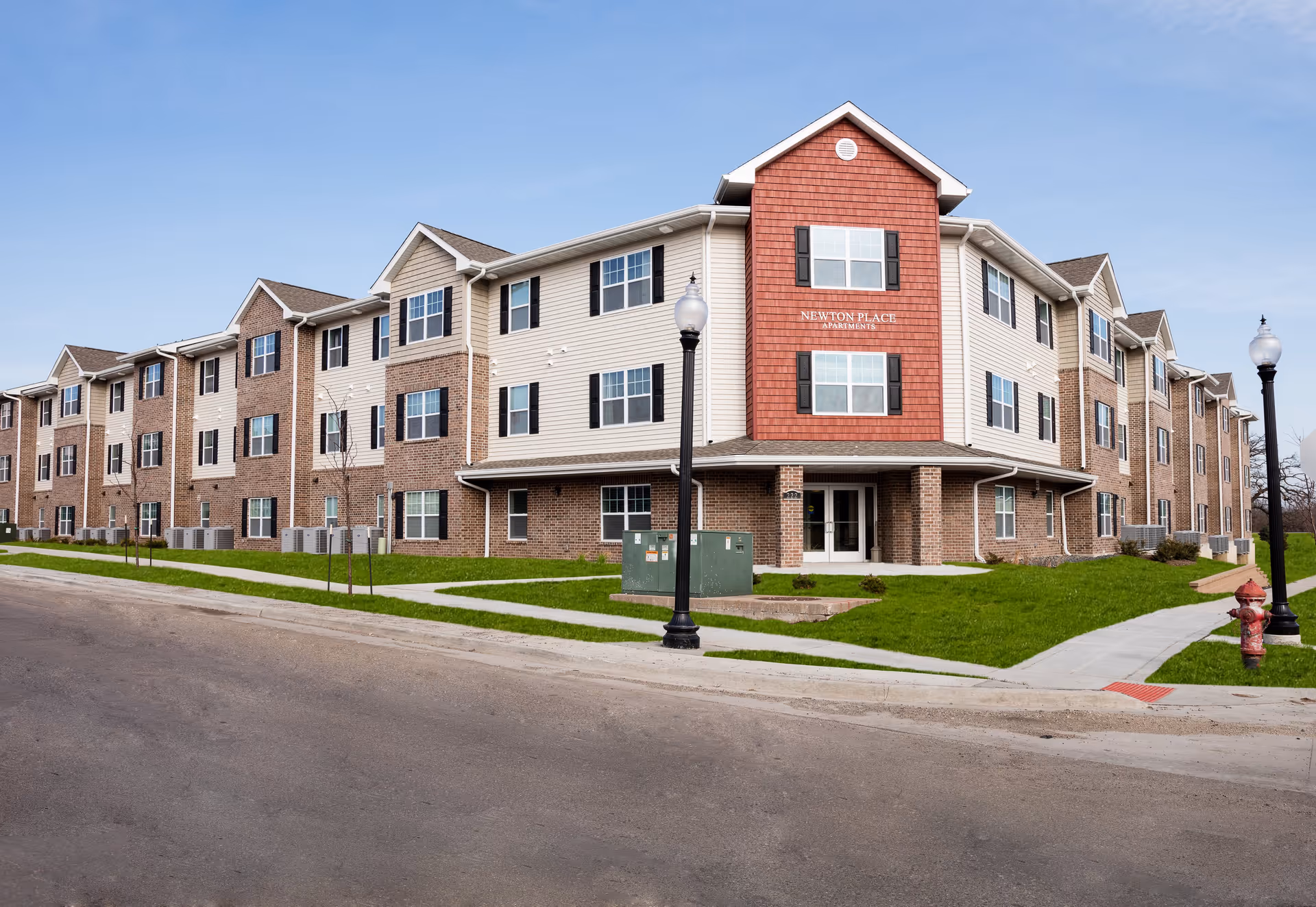 Three-story brick and siding apartment building labeled "Newton Place Apartments" with a lawn, sidewalk, street, lamp posts, and a fire hydrant in front.