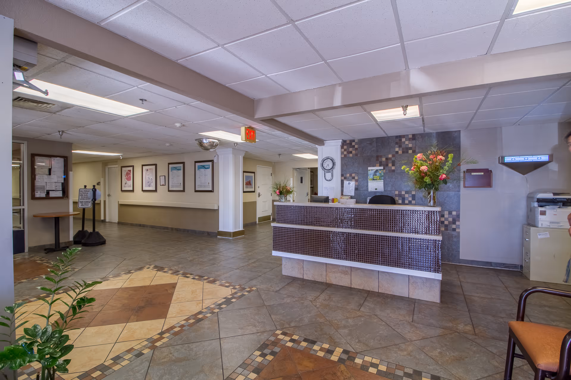 Reception area of Princeton Place facility featuring a front desk with a tiled front, a clock on the wall, floral arrangements, framed pictures on the walls, tiled flooring with decorative patterns, and a hallway leading to other rooms.