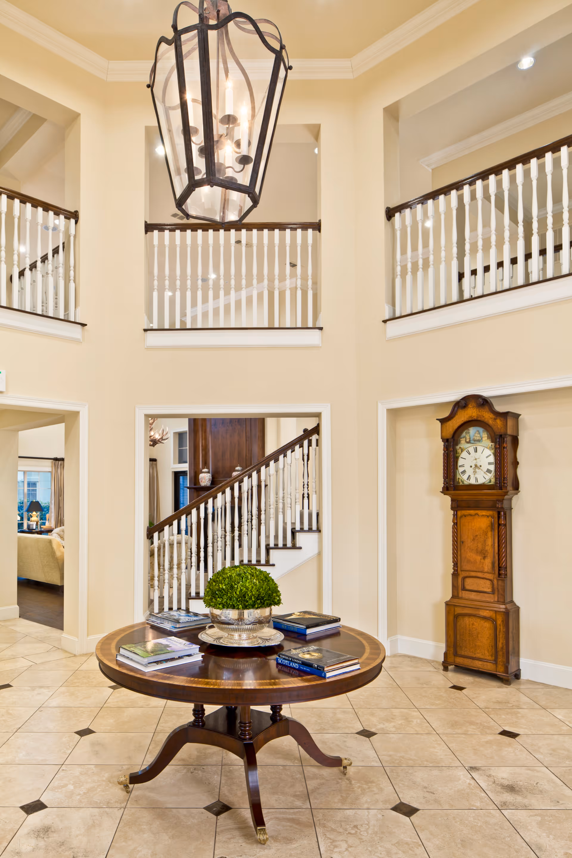 Spacious foyer with a large hanging lantern light fixture, a round wooden table with books and a decorative plant in the center, beige tiled floor, white railings on the upper level, and a tall wooden grandfather clock against the wall.