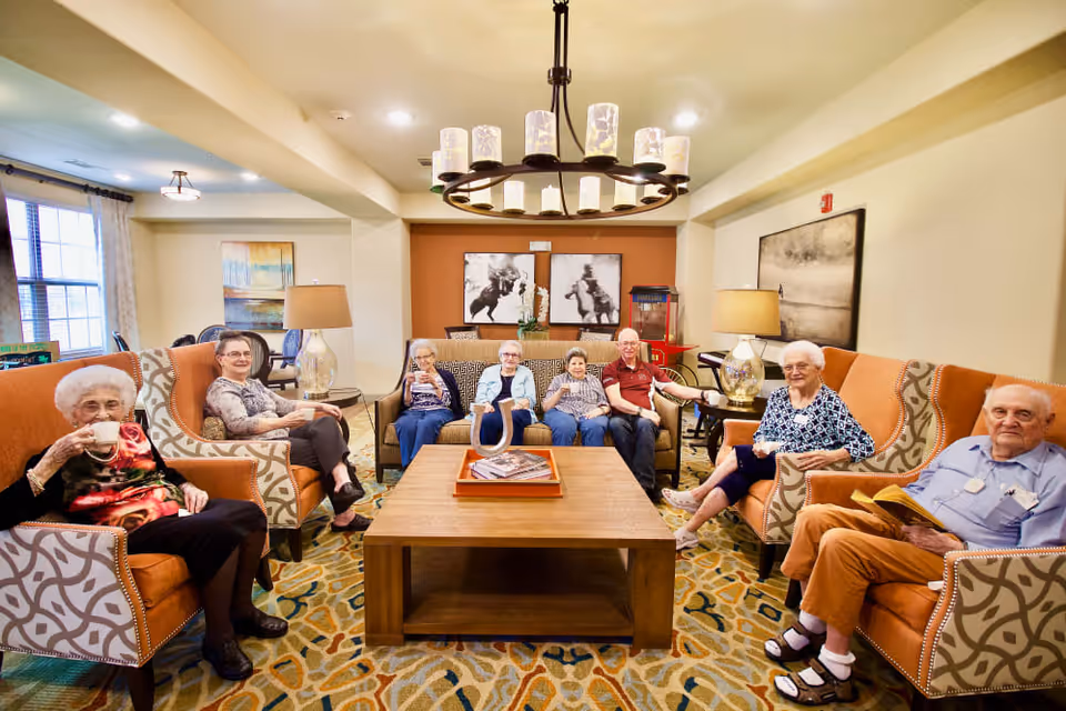 A group of elderly people sitting comfortably in a well-lit living room with patterned armchairs and a wooden coffee table. They appear relaxed, some holding cups and one person reading a book. The room has warm lighting, decorative lamps, and artwork on the walls.