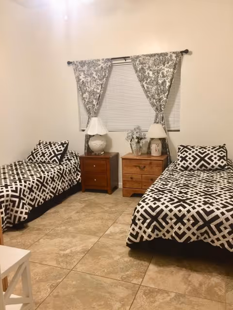 A bedroom with two twin beds featuring black and white geometric patterned bedding. Between the beds are two wooden nightstands, each with a lamp and decorative items. A window with white blinds and floral curtains is centered on the wall behind the nightstands.