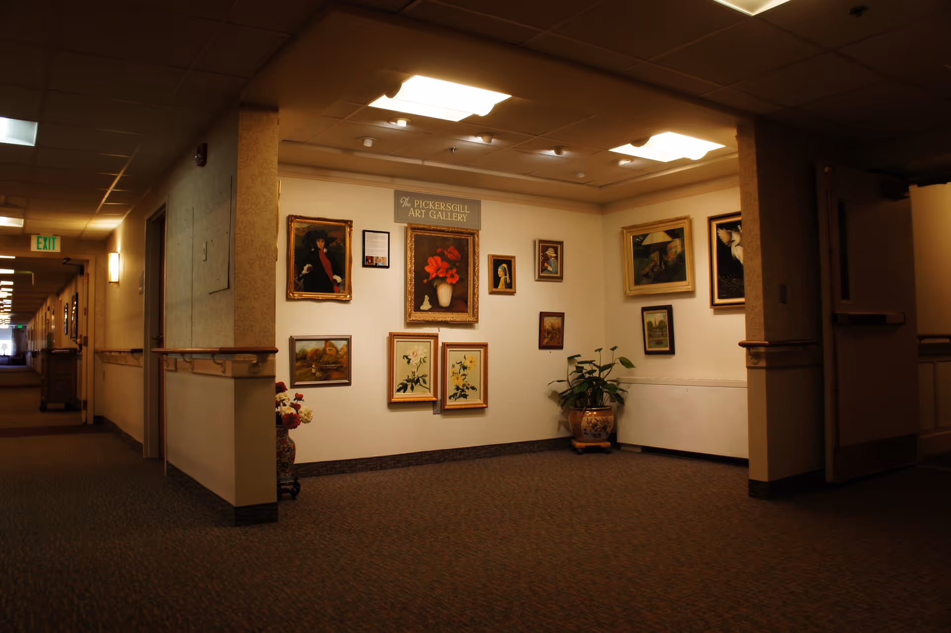 Interior hallway corner of Pickersgill Retirement Community featuring an art gallery wall with various framed paintings and a sign reading 'The Pickersgill Art Gallery'. There are two potted plants on the floor, one on each side of the gallery wall. The hallway extends to the left with doors and an exit sign visible.