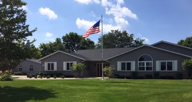 Single-story building with gray siding and multiple windows under a blue sky with scattered clouds. An American flag is flying on a flagpole in front of the building, which is surrounded by a well-maintained green lawn and some trees.